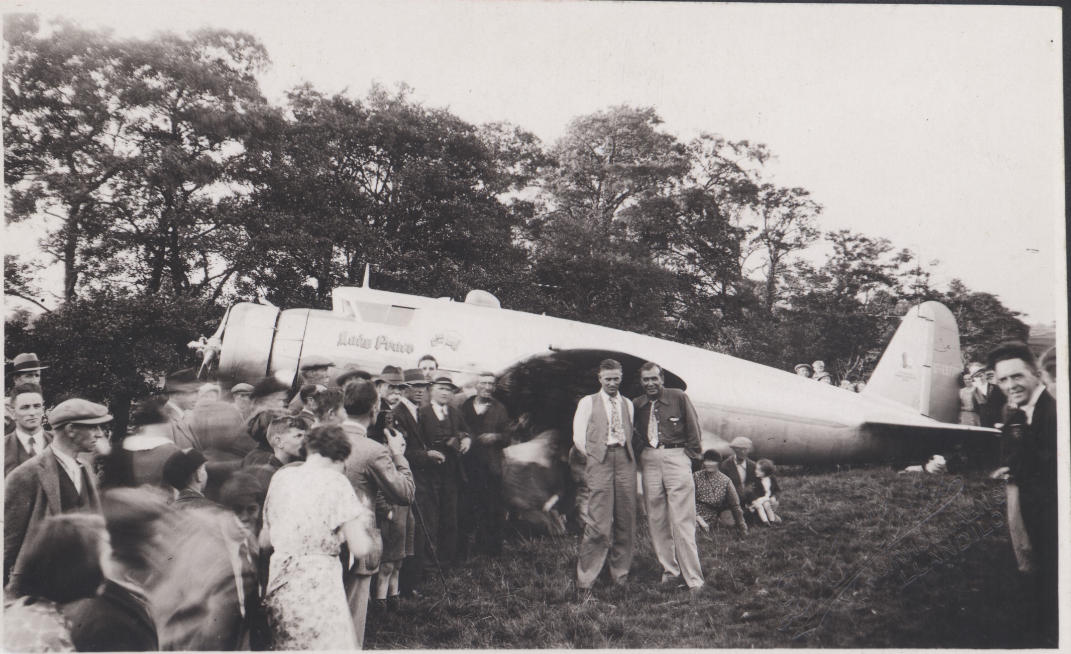 Black and white photograph of a crowd forming around an aeroplane (the Lady Peace), with two men front and centre.