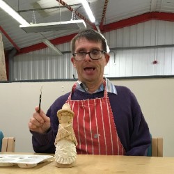 Carl with a paint brush in his hand getting ready to paint a pottery Welsh lady which is on the table in front of him.