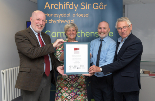 Pedwar oedolyn yn sefyll o flaen baner Archifdy Sir Gaerfyrddin, yn dal tystysgrif achredu.