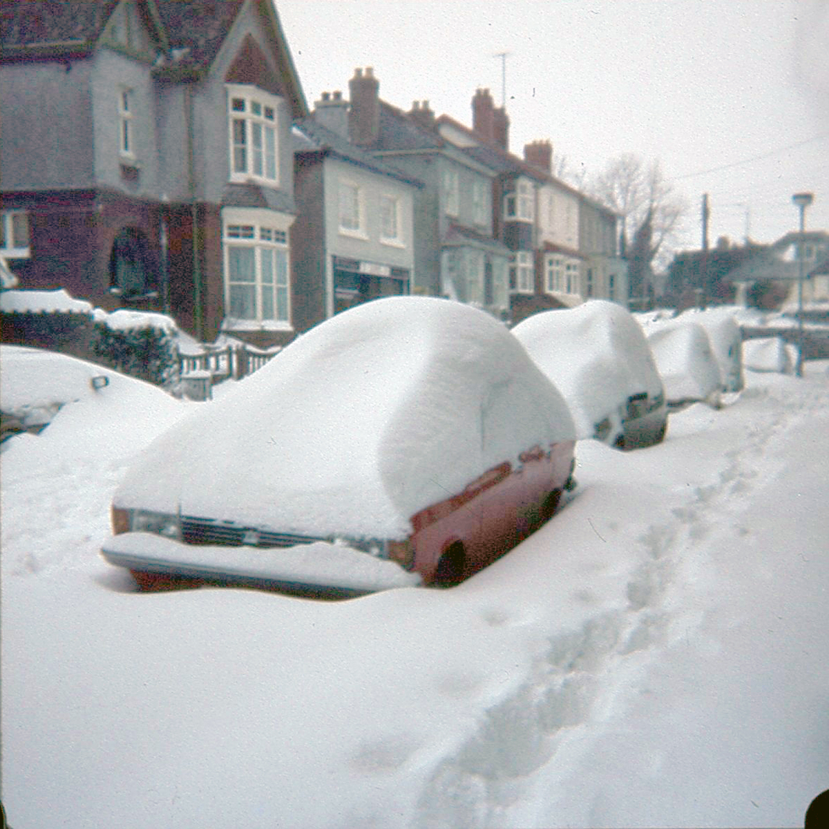 A red car at the forefront of the photograph covered in snow, followed by a line of cars seemingly parked outside a row of houses (also covered in snow).