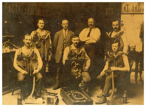 Sepia photograph of seven men - five suited up in old-fashioned breathing apparatuses, and two standing behind the group looking authoritative. Various mining equipment is spread over the floor.