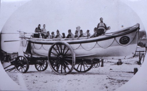Black and white photograph of a lifeboat atop a large set of wooden wheels containing a dozen crew wearing old-fashioned life vests.