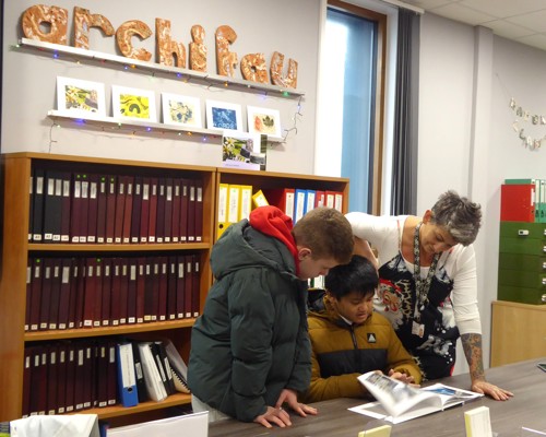 An adult looking on as two children flick through a book with ‘archifau’ spelt out in large ceramic letters in the background.